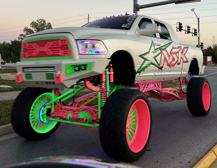Lifted truck with bright neon green and pink outrageous car mods driving on a city street at dusk.