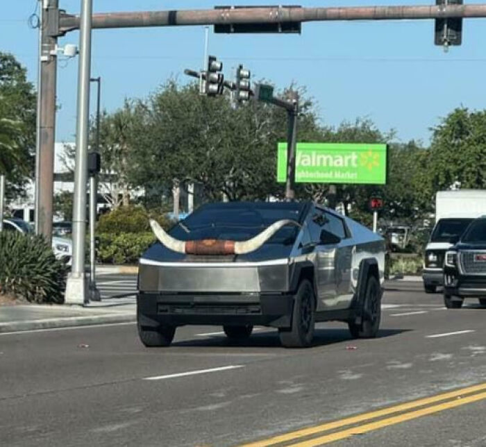 Silver Tesla Cybertruck with large bull horns attached to the front driving on a city street, an outrageous car mod example.