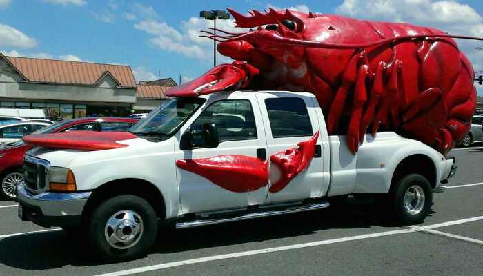 White truck with outrageous car mods featuring a giant red lobster attached to the roof and sides in a parking lot.