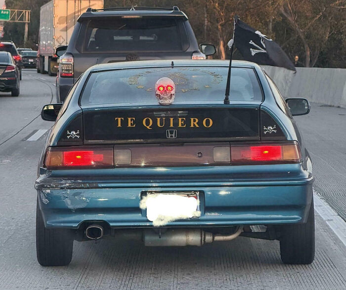 Blue car with skull decoration and pirate flag showcasing outrageous car mods on a busy highway at sunset.