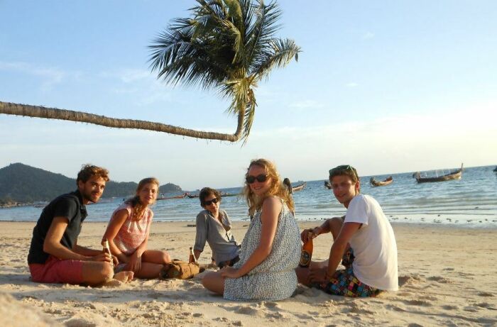 Group of friends sitting on beach under a bent palm tree in a funny nature moment captured by people snapping a pic.
