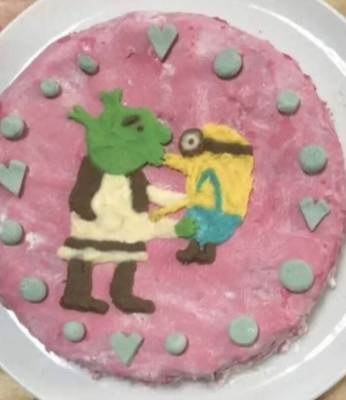 Cursed food cake with poorly made green ogre and yellow cartoon character decoration on pink frosting.