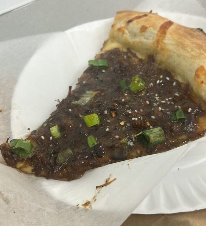 Slice of cursed food pizza topped with unappetizing dark sauce, green onions, and sesame seeds on a white paper plate
