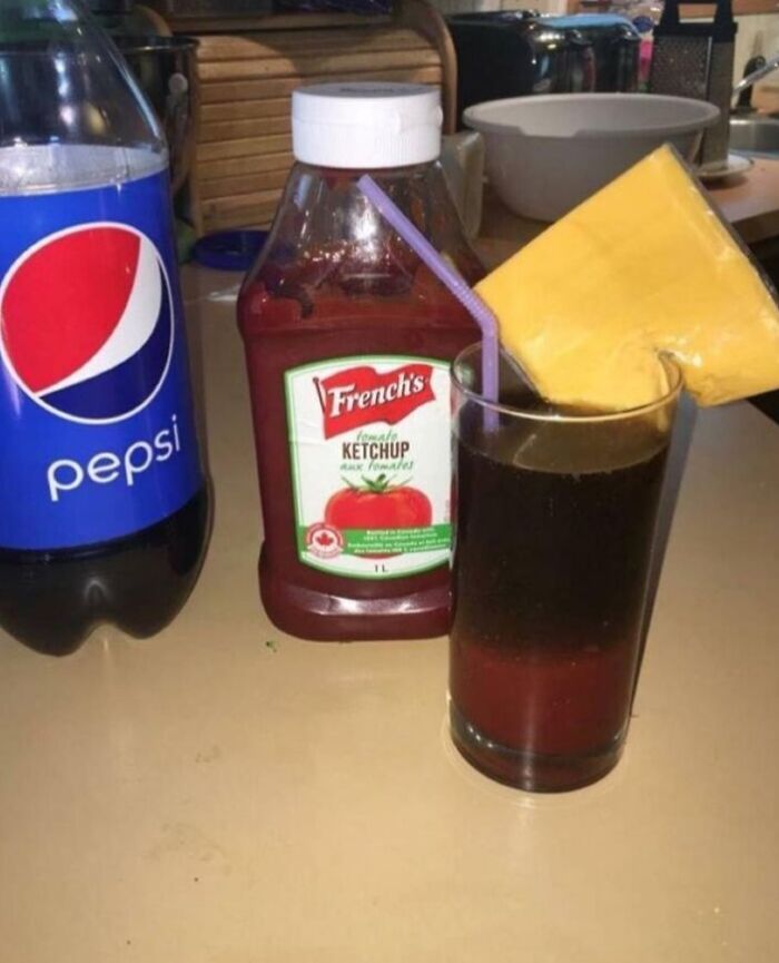 Glass filled with soda, a slice of processed cheese on the rim, bottle of ketchup and Pepsi bottle in the background, cursed food picture.