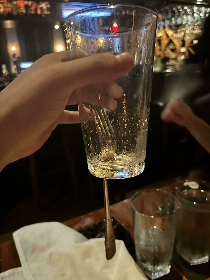 Hand holding a glass with a fork seemingly suspended inside, a mildly interesting visual shared at a bar.