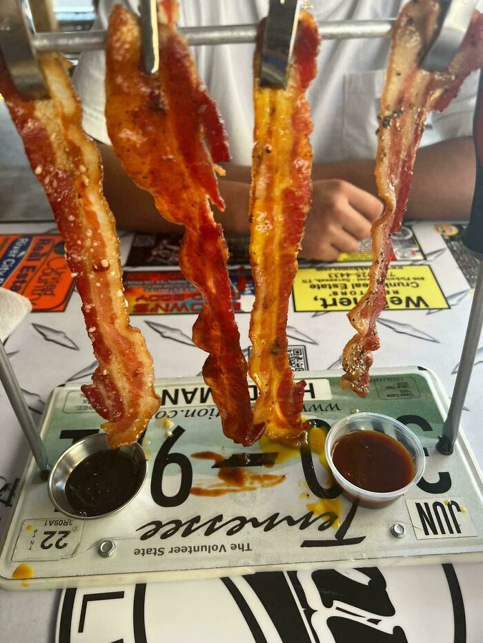 Strips of crispy bacon served hanging on a metal rack with dipping sauces on a license plate-style tray.