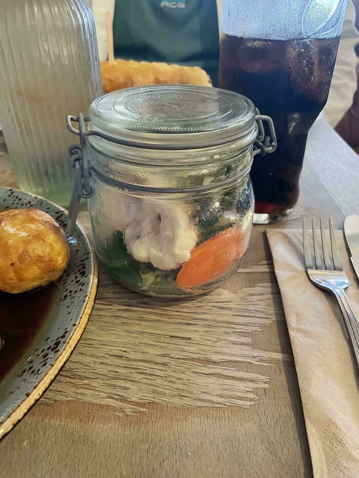 Unholy meal served with vegetables in a glass jar alongside a drink and bread roll on a wooden table.