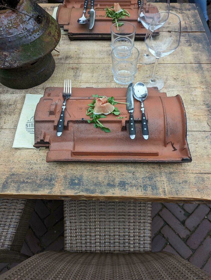 Unholy meal presentation with small salad served on a rooftop tile plate with cutlery and glassware on wooden table.