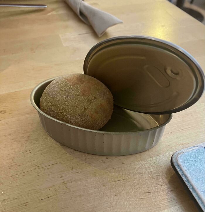 Unholy meal presentation with a bread roll served inside an open empty tin can on a wooden table.