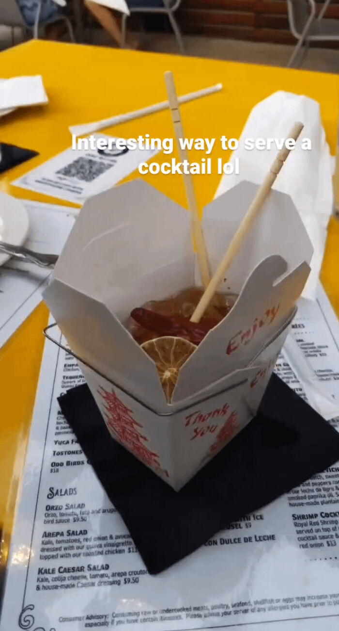 Cocktail served in an unusual way inside a Chinese takeout box with chopsticks and dried citrus garnish on a menu table.