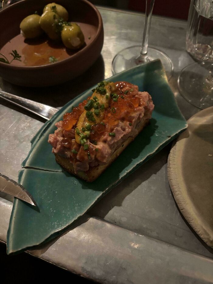 Unholy meal served on a broken ceramic plate with fish roe and garnish on a metal table at a dimly lit restaurant.