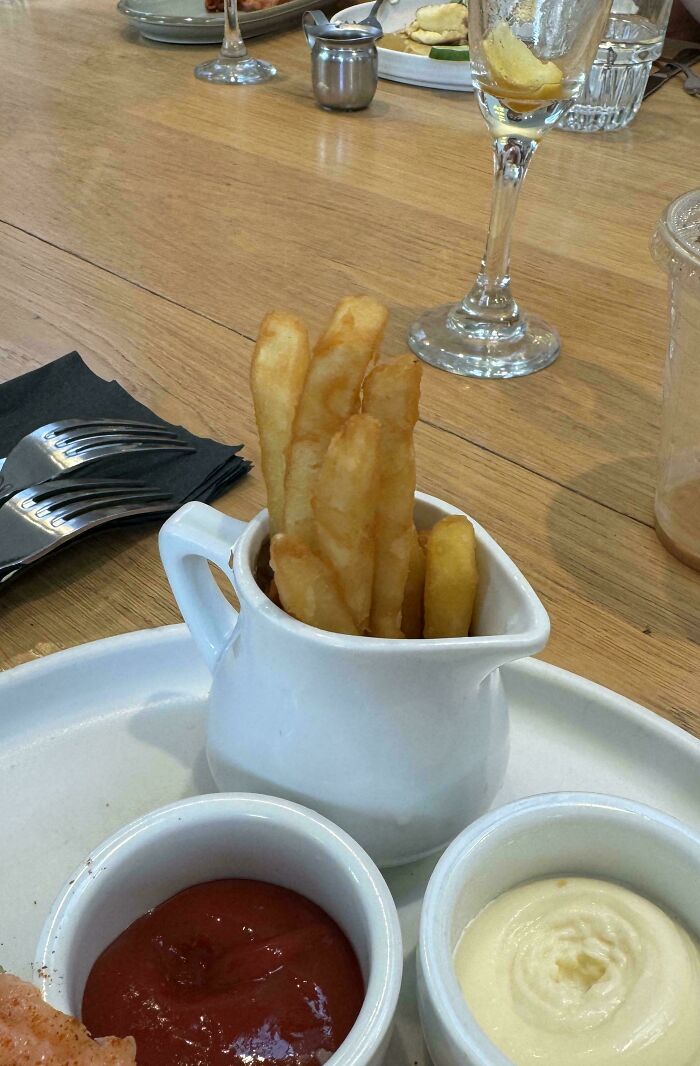 Fries served inside a small white pitcher alongside ketchup and mayonnaise dips on a wooden table.