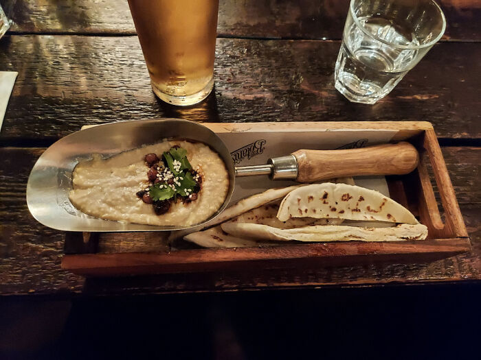 Metal scoop filled with dip and flatbread slices served on a wooden tray, an example of meals served in unholy ways.