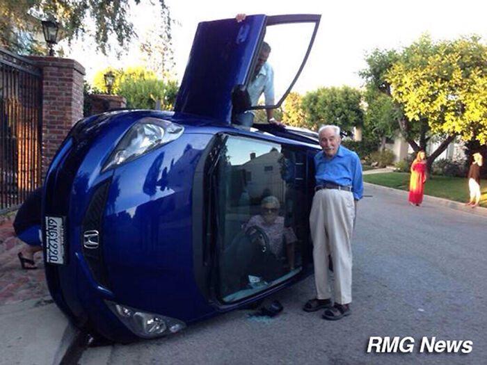 Elderly man standing beside a flipped blue car, looking calm and suspiciously unbothered in a suburban street.