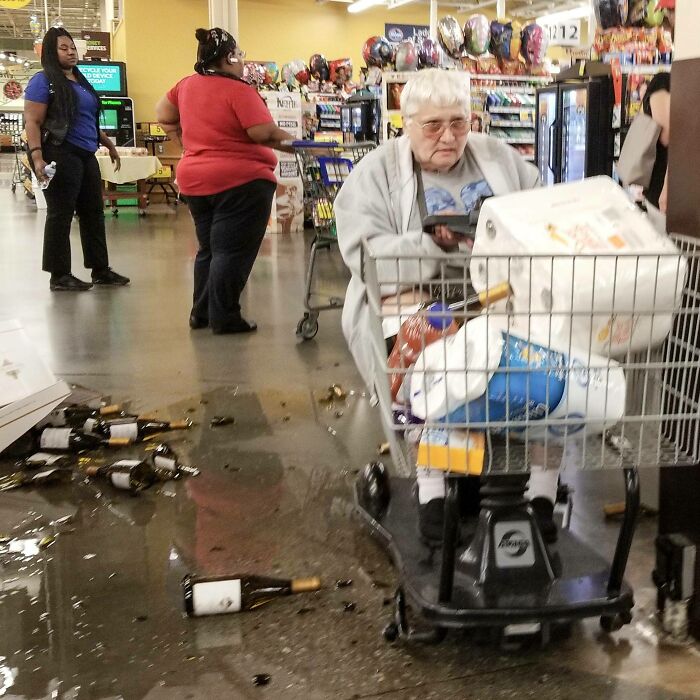Elderly woman on mobility cart shopping, surrounded by shattered wine bottles spilled on supermarket floor, looking unbothered.