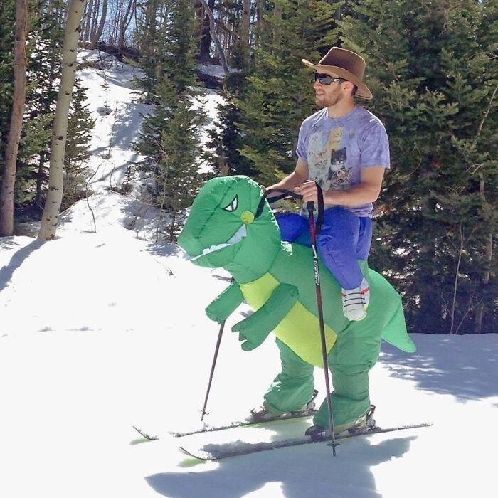 Man skiing in snowy forest wearing inflatable dinosaur costume, showing suspiciously unbothered attitude outdoors.