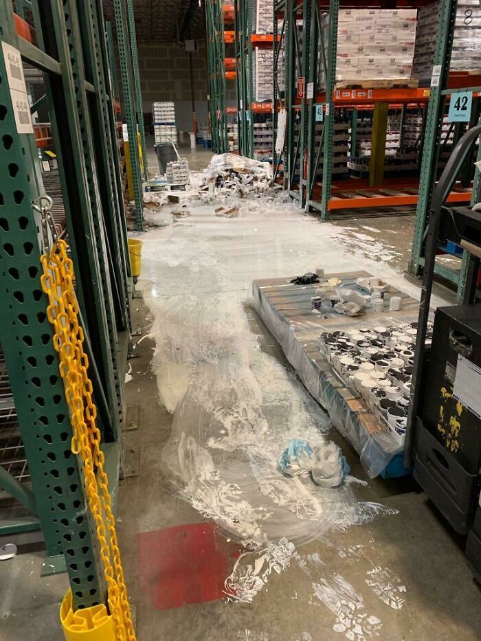 Warehouse work fail showing spilled white paint or powder across the floor and damaged stacked products on pallets.