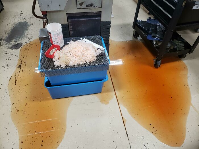 Rusty liquid spilled on factory floor beside stacked blue bins and scattered debris, showcasing epic work fail cleanup challenge.