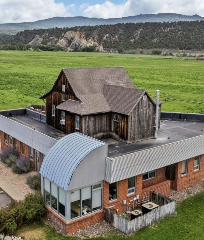 Unhinged real estate listing showing a rustic wooden house oddly placed atop a modern brick building in a rural landscape.