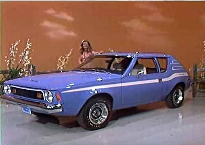 Vintage blue compact car on display indoors with a woman standing behind, showcasing unique vintage things collection.