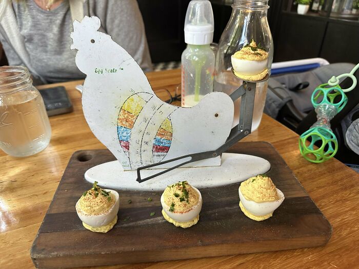 Unholy meal presentation of deviled eggs served in an unusual chicken-shaped metal holder on a wooden board.