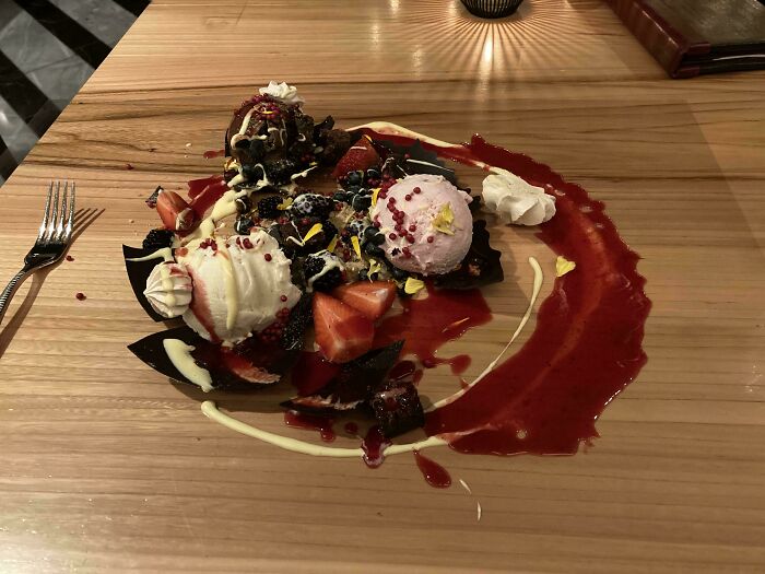 Dessert served messily with ice cream, strawberries, sauce, and whipped cream on a wooden table, showcasing unholy meal presentation.