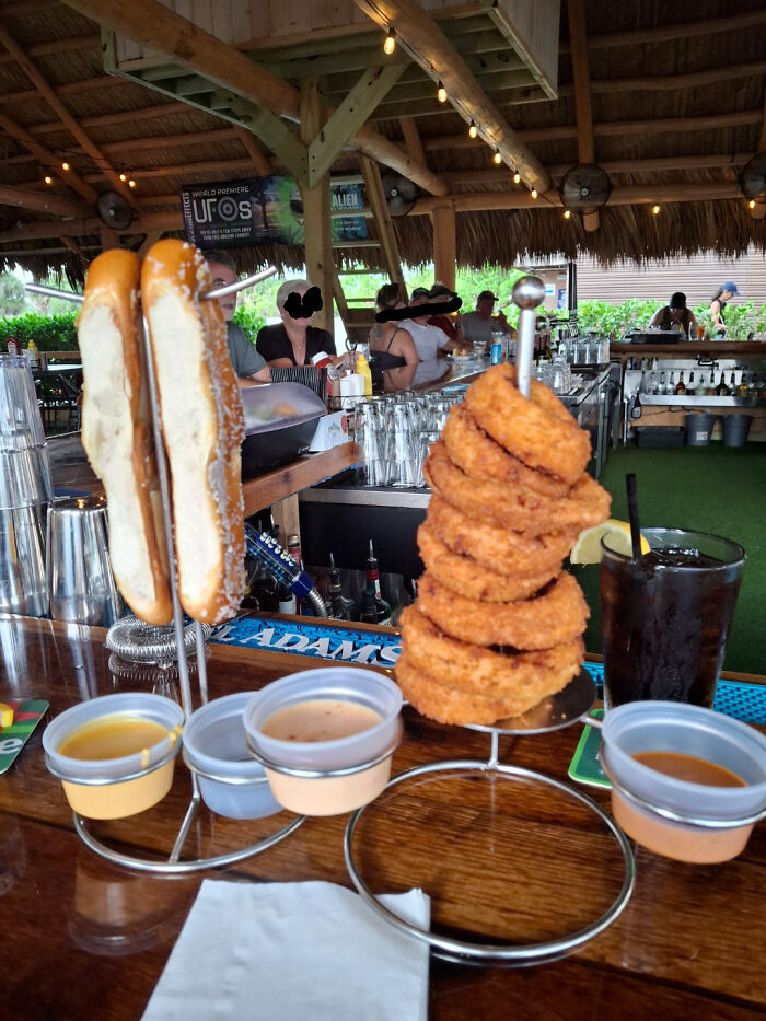 Unholy meal serving with onion rings stacked on a skewer and pretzel buns on metal holders with dipping sauces at a bar.