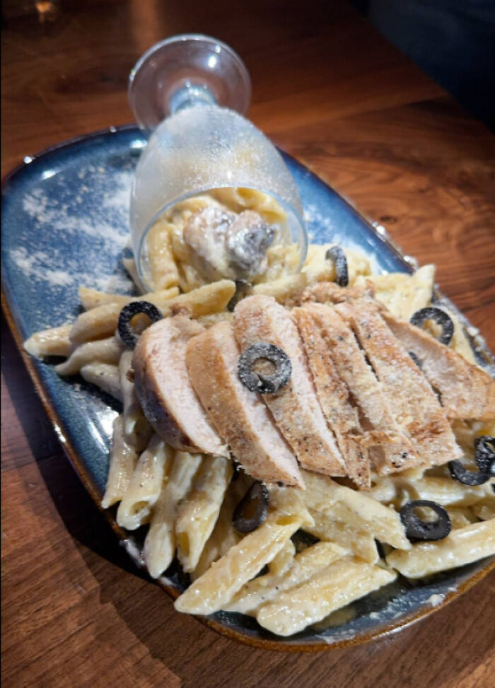 Pasta with sliced chicken and black olives served with upside-down glass on a plate, showcasing unholy meal presentation.