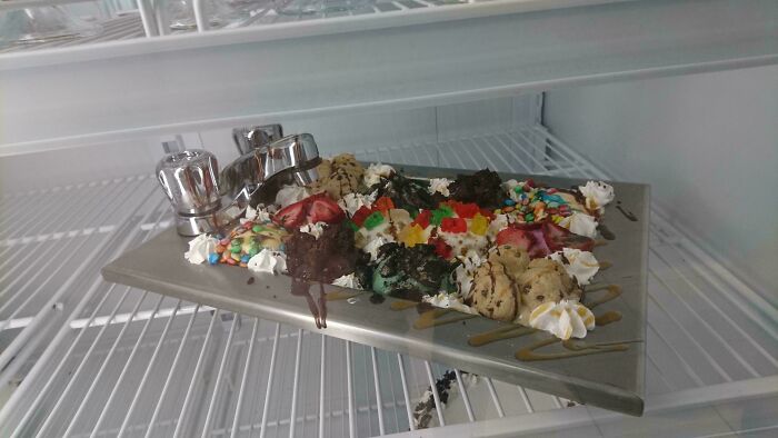 Unholy meal presentation of dessert with ice cream, whipped cream, gummies, and cookies served on a metal tray inside a fridge.