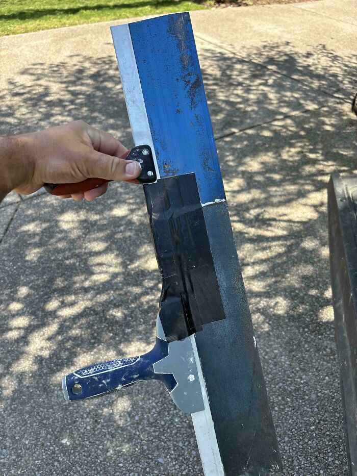 Hand holding a duct-taped tool made from a paint scraper and metal blade showcasing redneck engineering creativity outdoors.