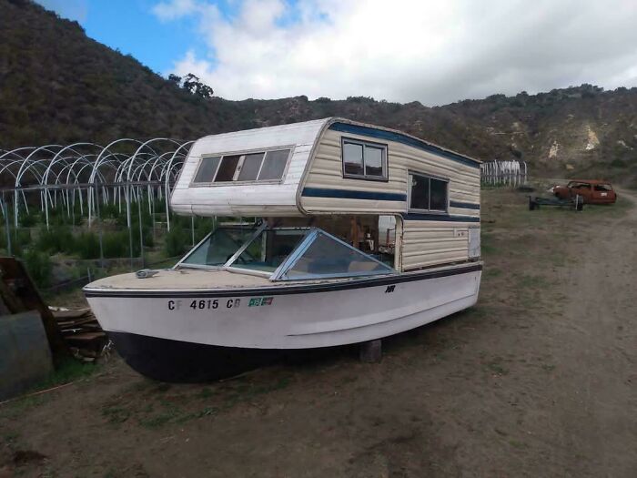 Boat converted into a camper showcasing creative redneck engineering in a rural outdoor setting with hills in the background.