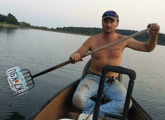 Man in a boat using a makeshift paddle made from a pitchfork and license plate, showcasing redneck engineering creativity.