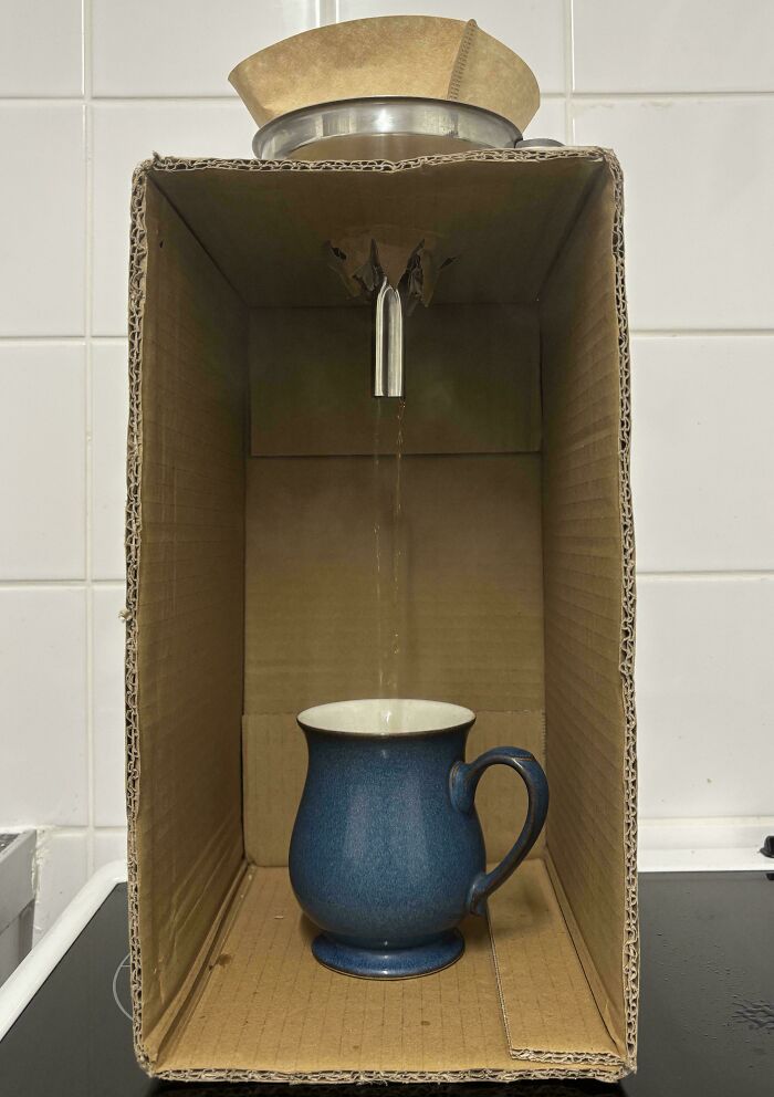 Improvised cardboard coffee drip setup with stream pouring into blue mug demonstrating redneck engineering creativity.