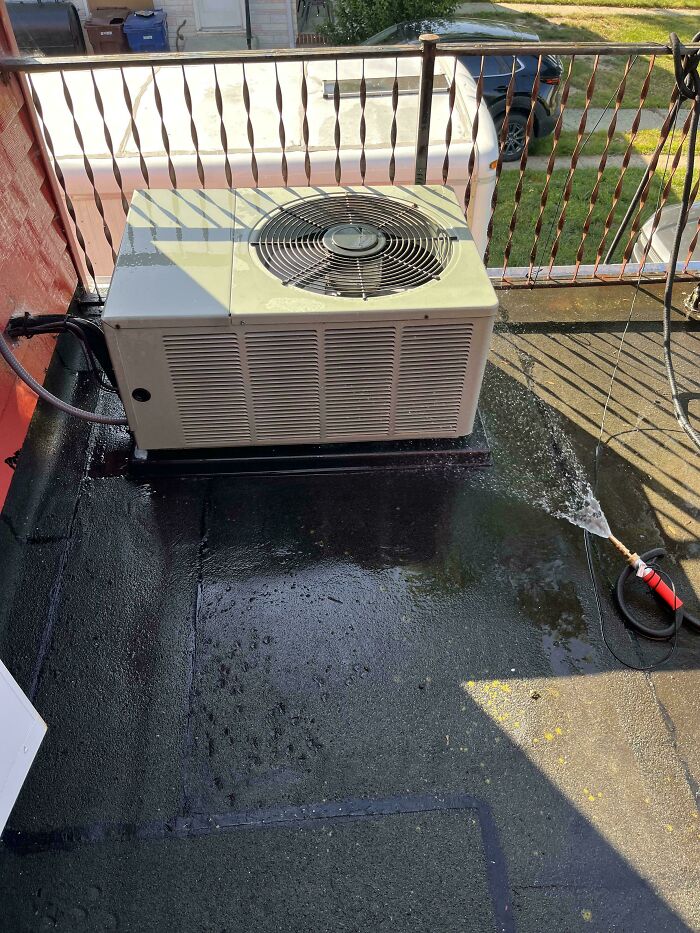 Outdoor air conditioning unit on black rooftop with water hose spraying nearby, showcasing redneck engineering creativity.