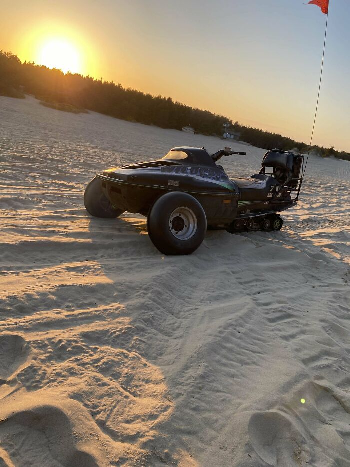 Off-road vehicle with large rear tire and tracks creatively modified for sand terrain at sunset in a desert setting.