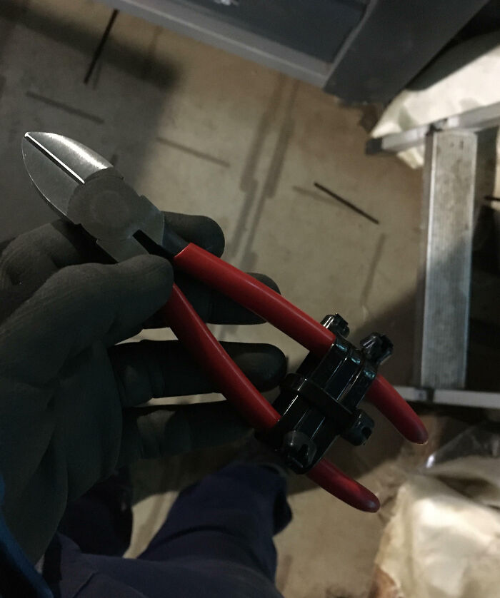 Hand wearing a glove holding pliers with red handles showing a unique work fail involving tool modification indoors.