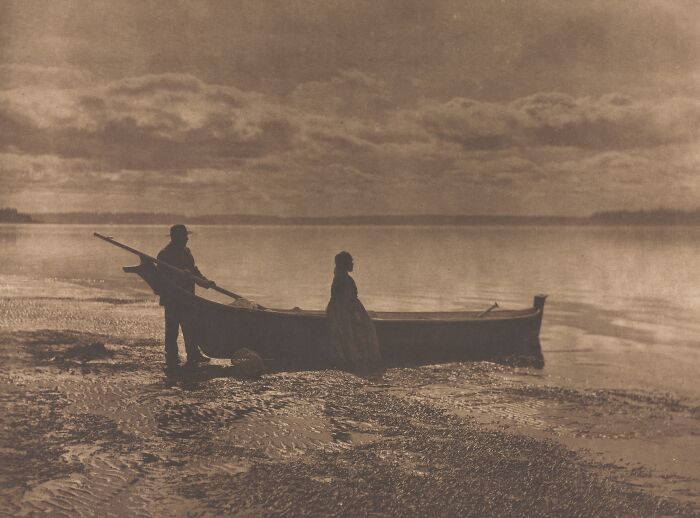 Two Native Americans by a canoe at a calm lakeshore during sunset, Edward S. Curtis photo preserving history.