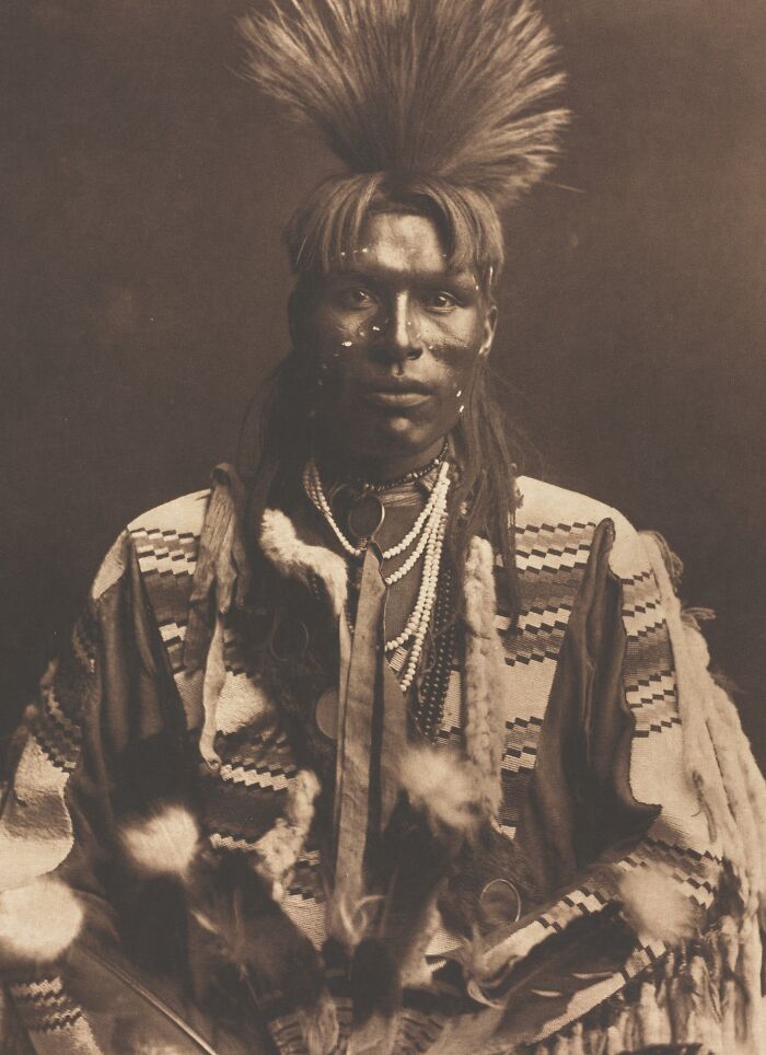 Native American man in traditional dress with feathered headdress and face paint in a powerful Edward S. Curtis photo.