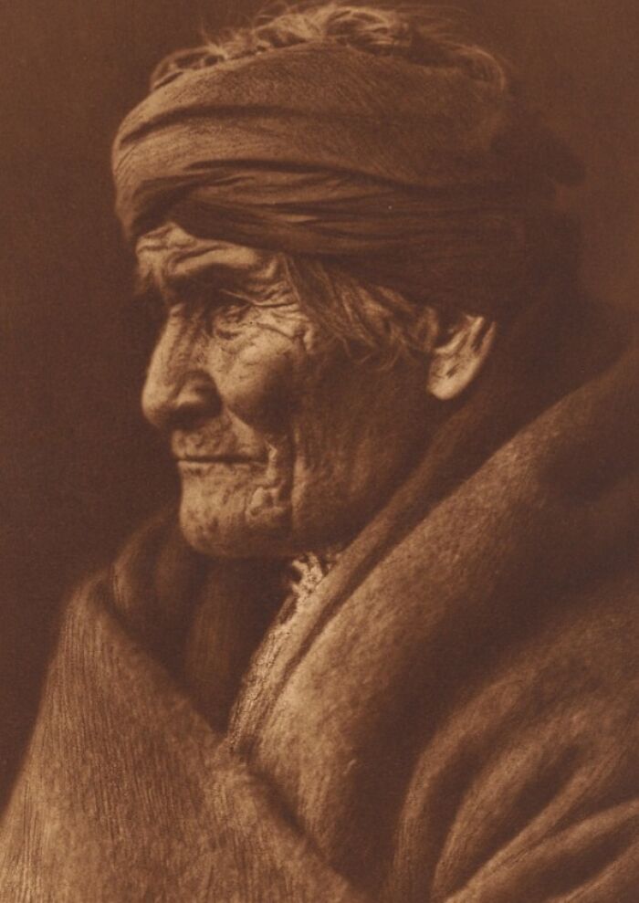 Sepia-toned Edward S. Curtis photo of an elderly Native American wrapped in a blanket, showcasing detailed facial wrinkles.
