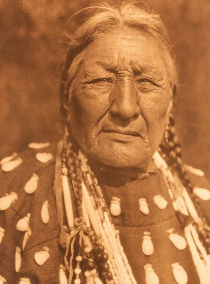 Elderly Native American woman with braided hair wearing traditional clothing in a powerful Edward S. Curtis photo.
