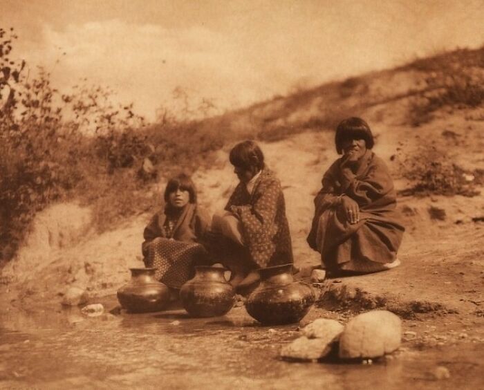 Three Native American children in traditional clothing sitting by a water source with pots in an Edward S. Curtis photo.