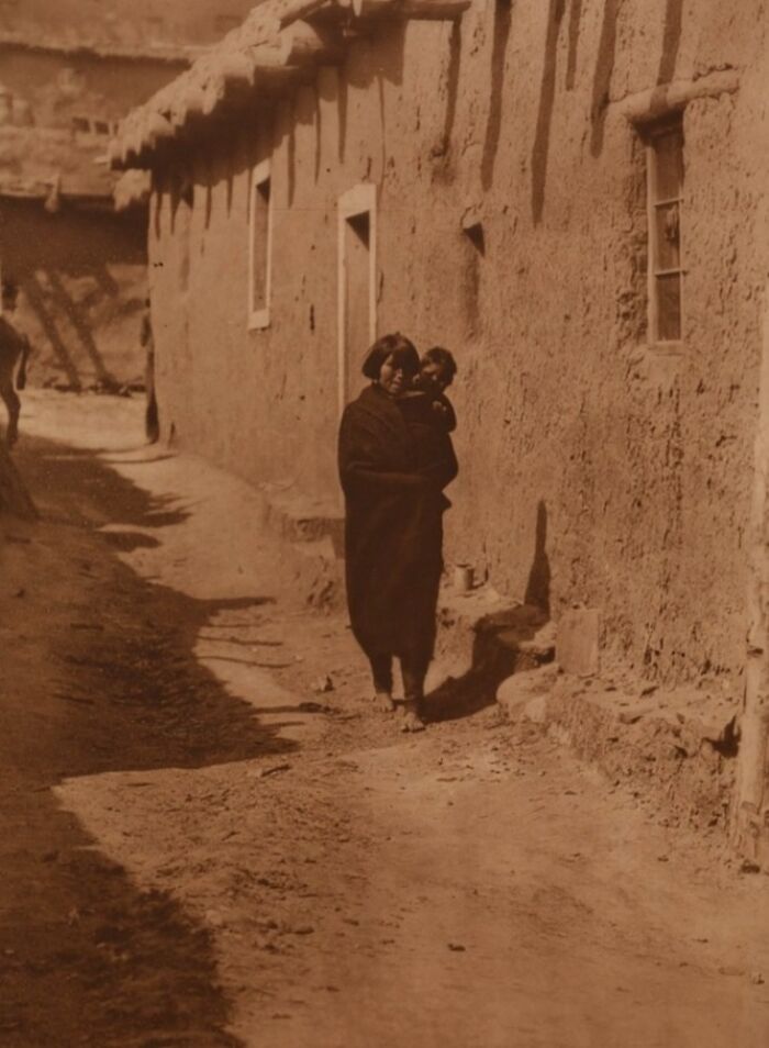 Native American woman holding child walking along adobe building in historic Edward S. Curtis photo preserving Native American history.