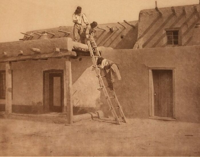 Three Native American men using a traditional ladder on an adobe building roof in a powerful Edward S. Curtis photo.