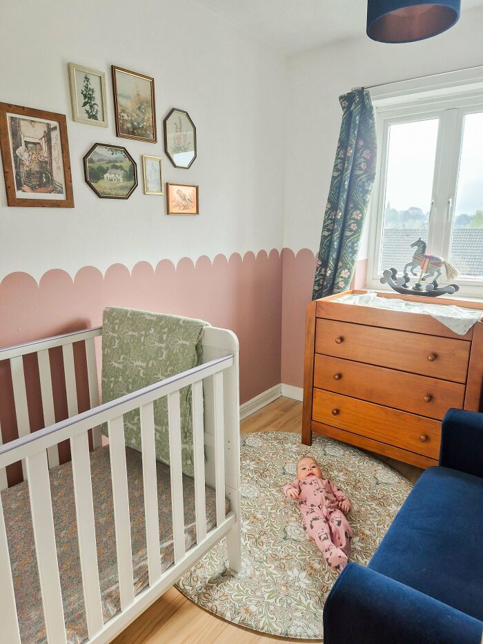 Baby lying on patterned rug in a revamped living space with a white crib, wooden dresser, and floral curtains.