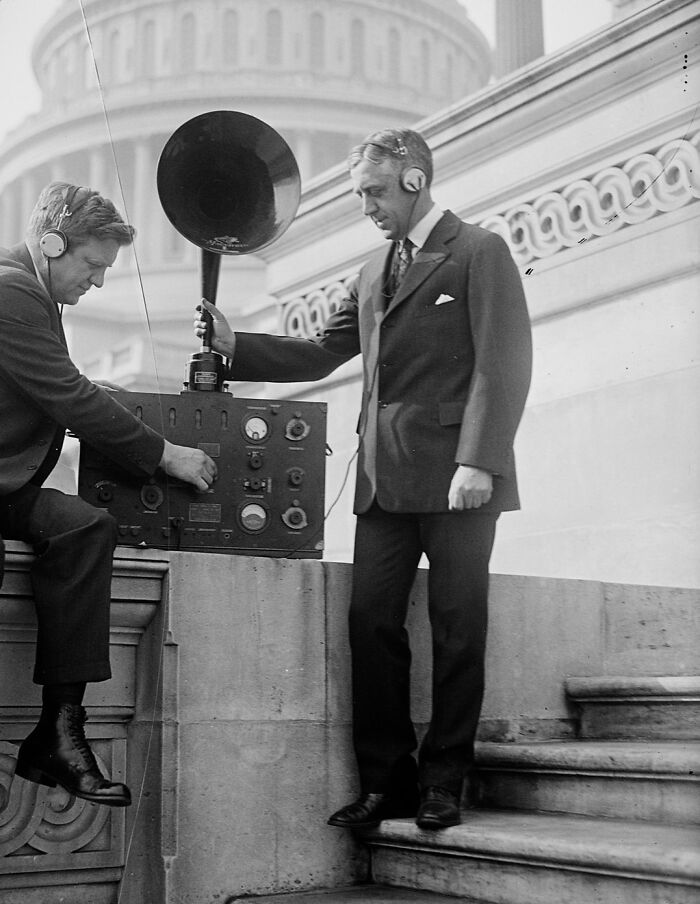 Two men with headphones operating vintage radio equipment outside a historic Washington, D.C. building in the 1920s.