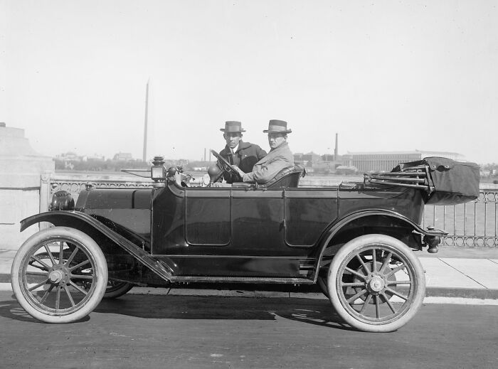 Two men wearing hats seated in a vintage car in Washington, D.C., with city landmarks visible in the background.