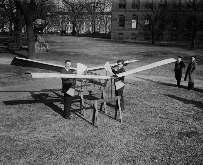 Two men in 1920s Washington, D.C., working on a large vintage model aircraft outdoors on a grassy lawn.