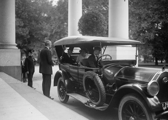 Vintage 1920s car with passengers and men standing near columns in a historical Washington, D.C. street scene.