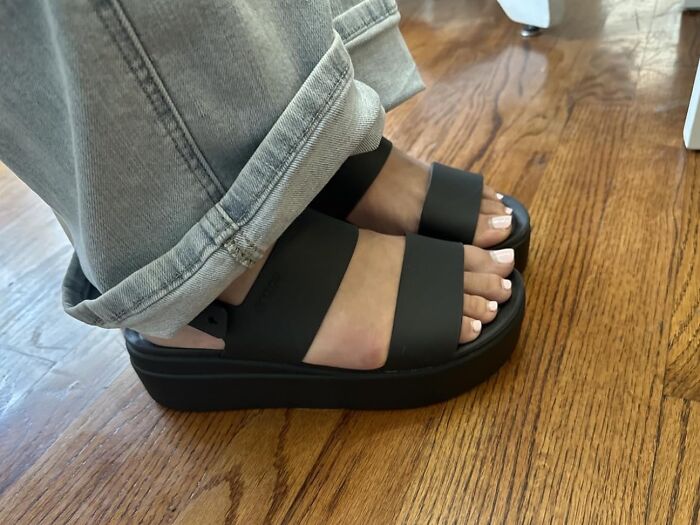 Close-up of black platform sandals on feet with painted nails, paired with casual rolled-up denim jeans on wooden floor, travel essentials.