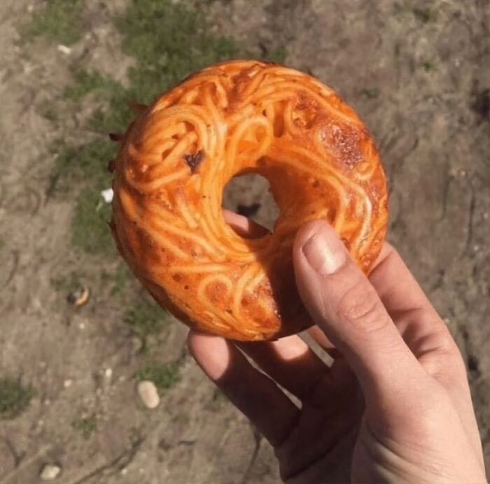 Hand holding a cursed food item resembling a bagel with baked spaghetti noodles embedded, creating a strange food appearance